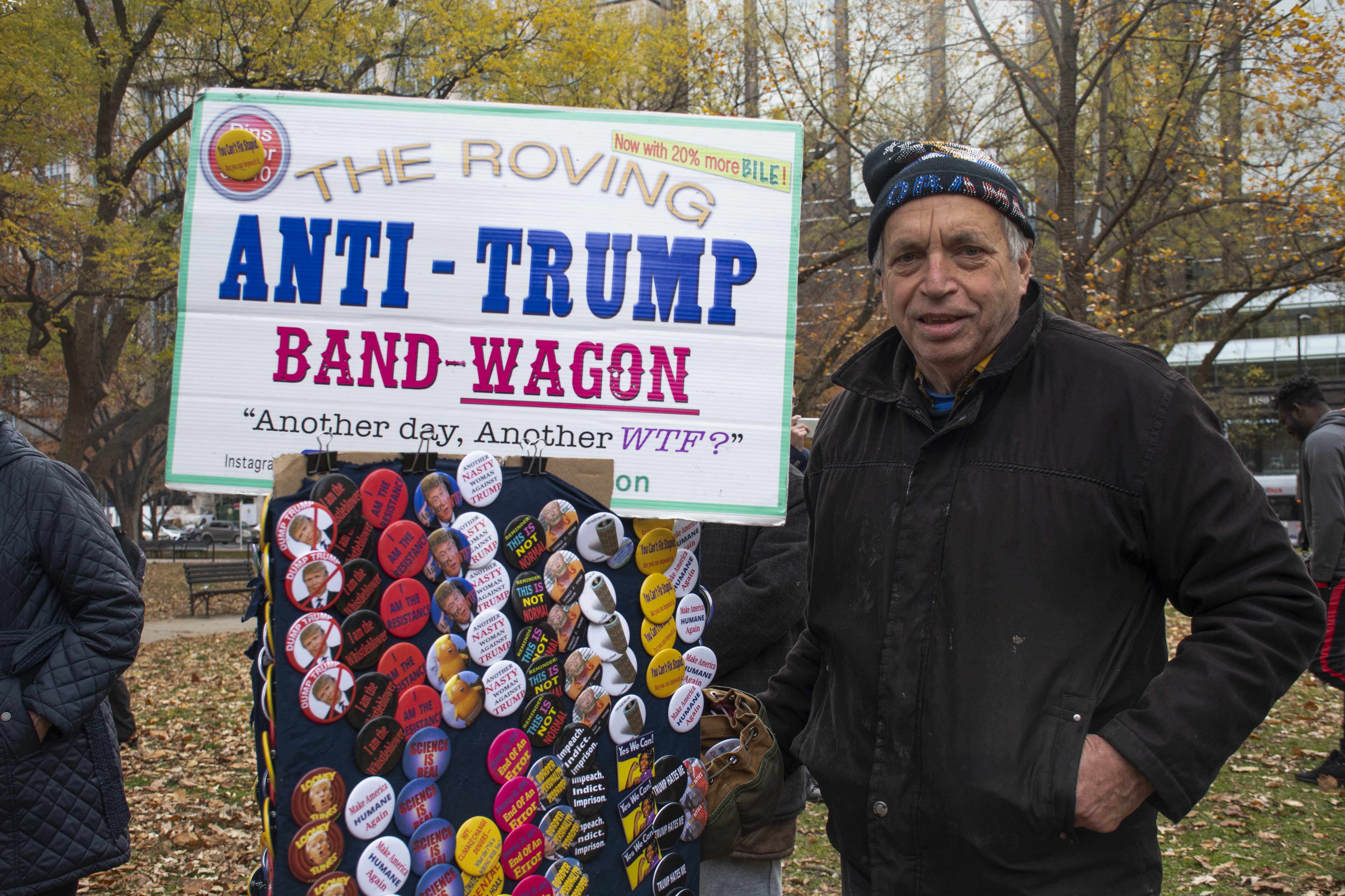 A man poses by a display of pins, with a sign that reads: The roving Anti-Trump Band-Wagon. Another day, Another WTF