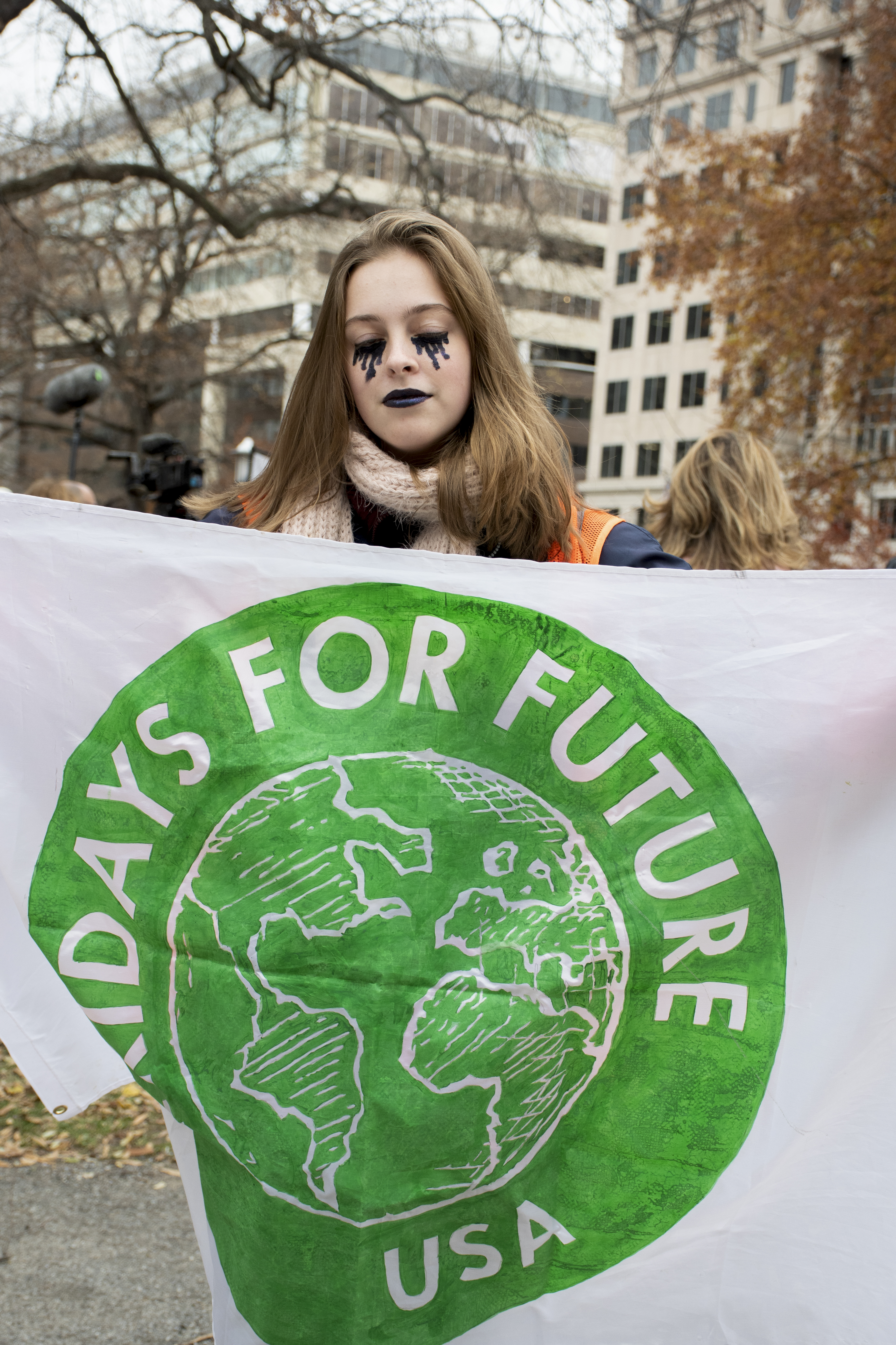 Teenage girl, eyes closed and with black tears painted onto her cheeks, holds up a sheet that says: Fridays for Future USA