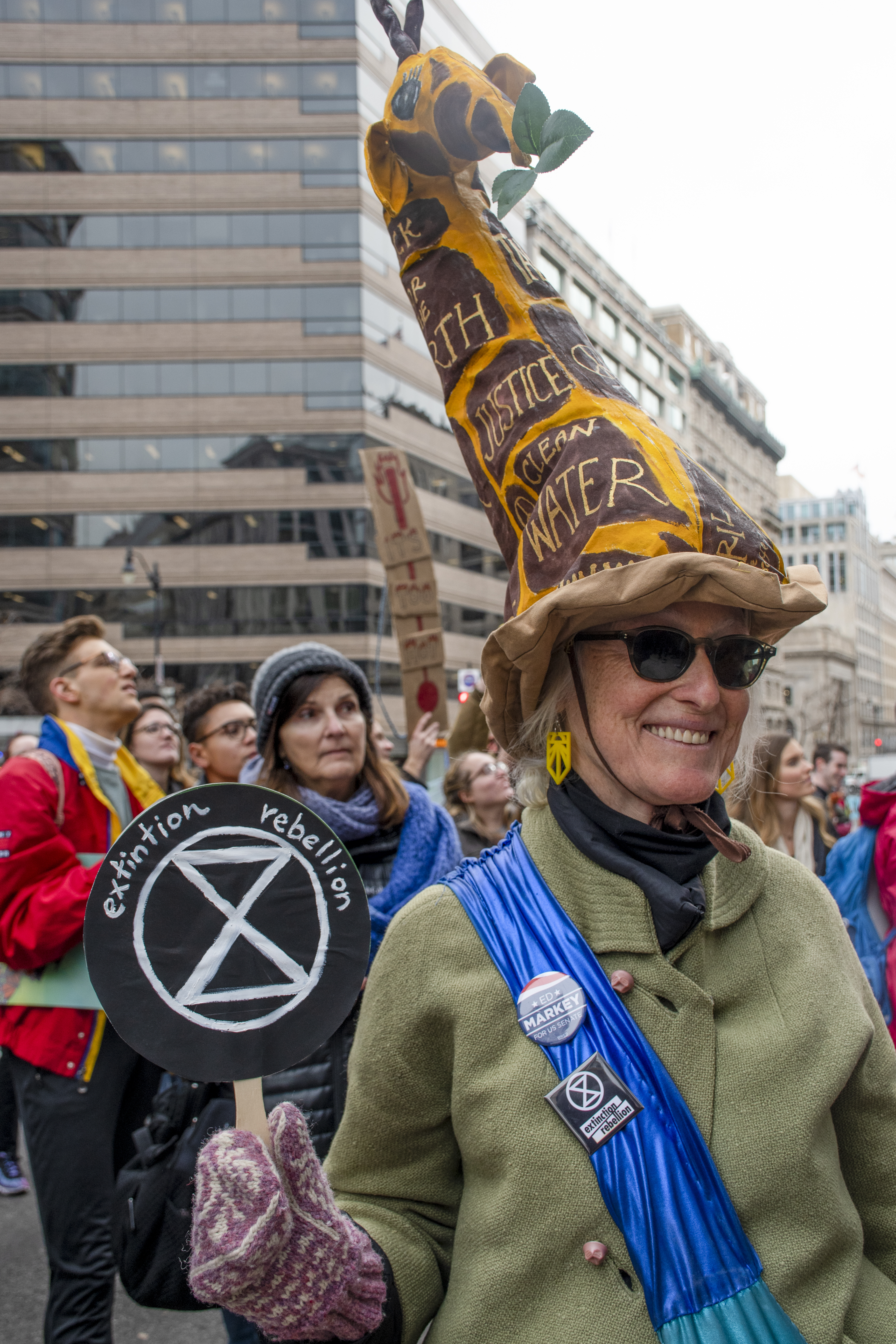 A woman with a hat shaped like a giraffe on her head hols up a small sign that says: Extinction rebellion