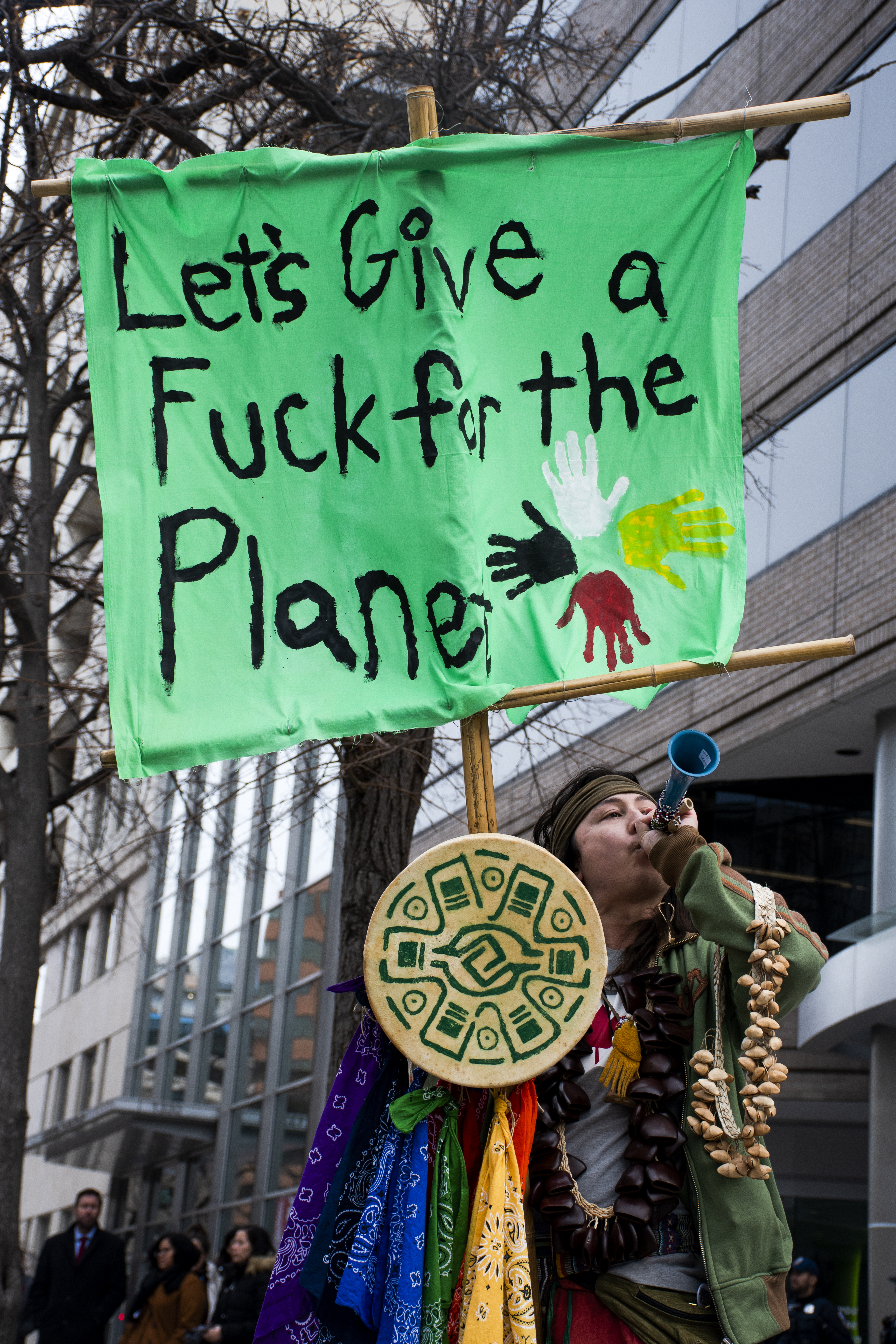A man in Native American dress blows into an intrument and holds up sign that reads: Let's give a fuck for the planet