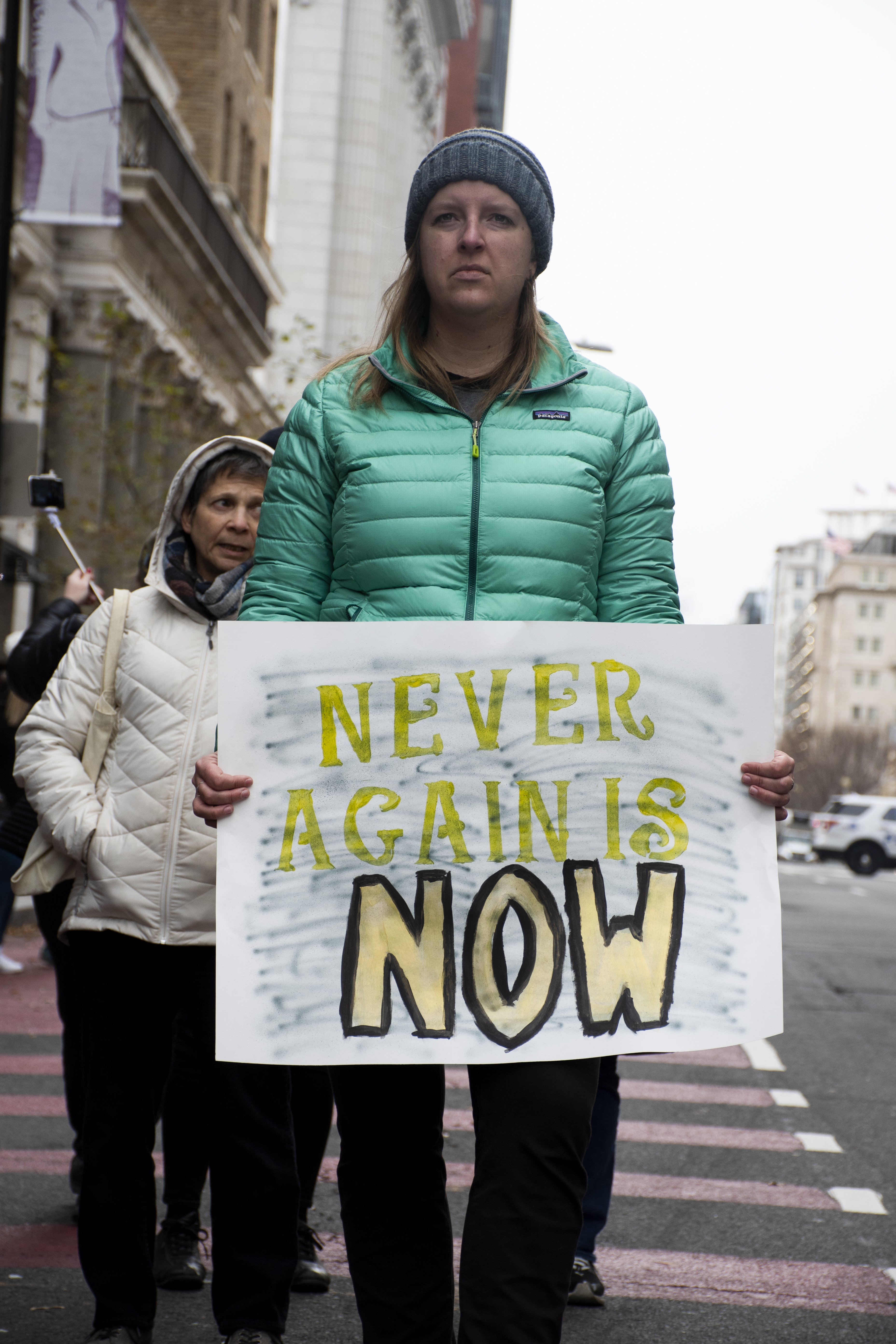 Woman in teal winter coat holds up sign that reads: Never again is now