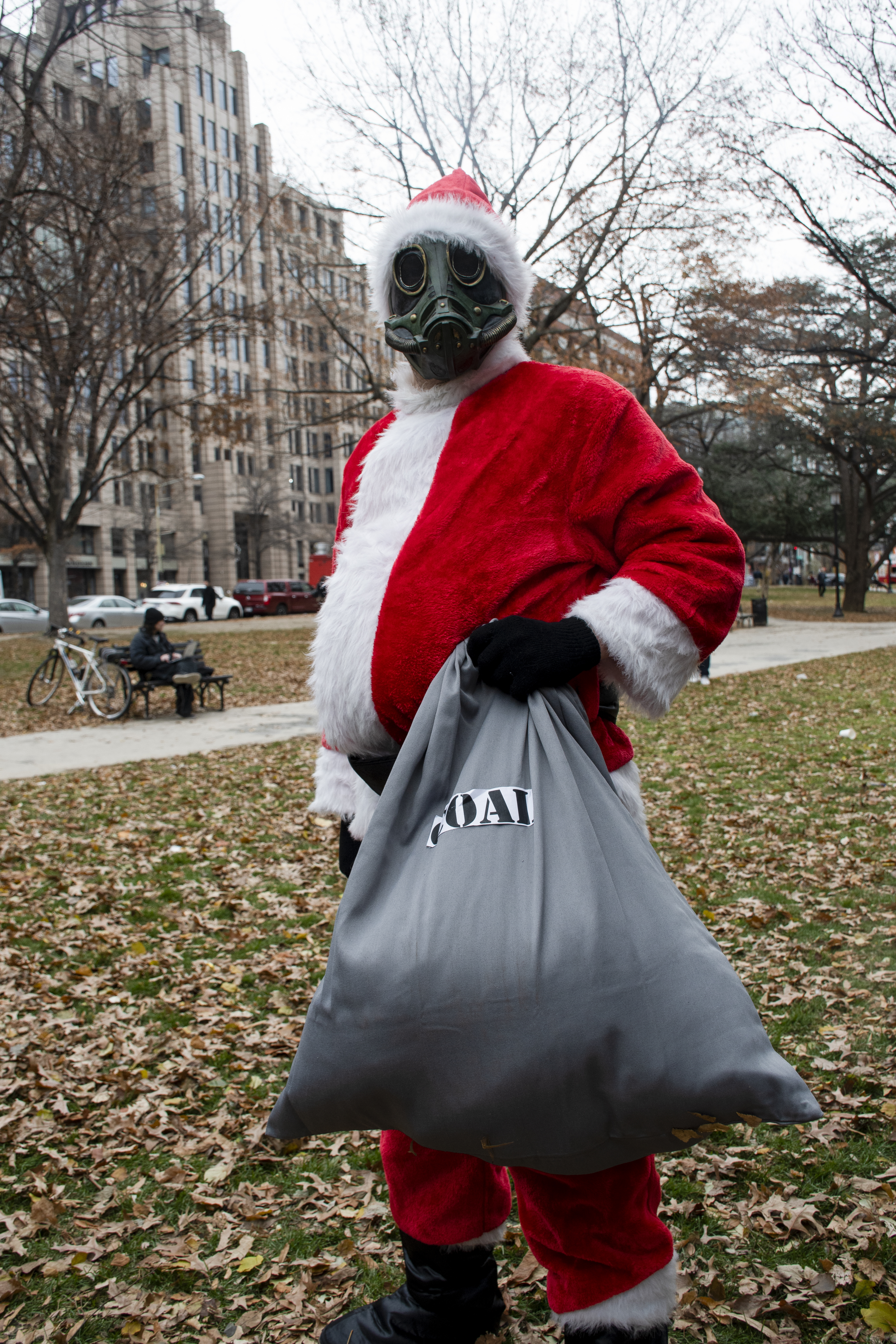 A man dressed as Santa Claus wears a gas mask and holds up a bag labeled as coal