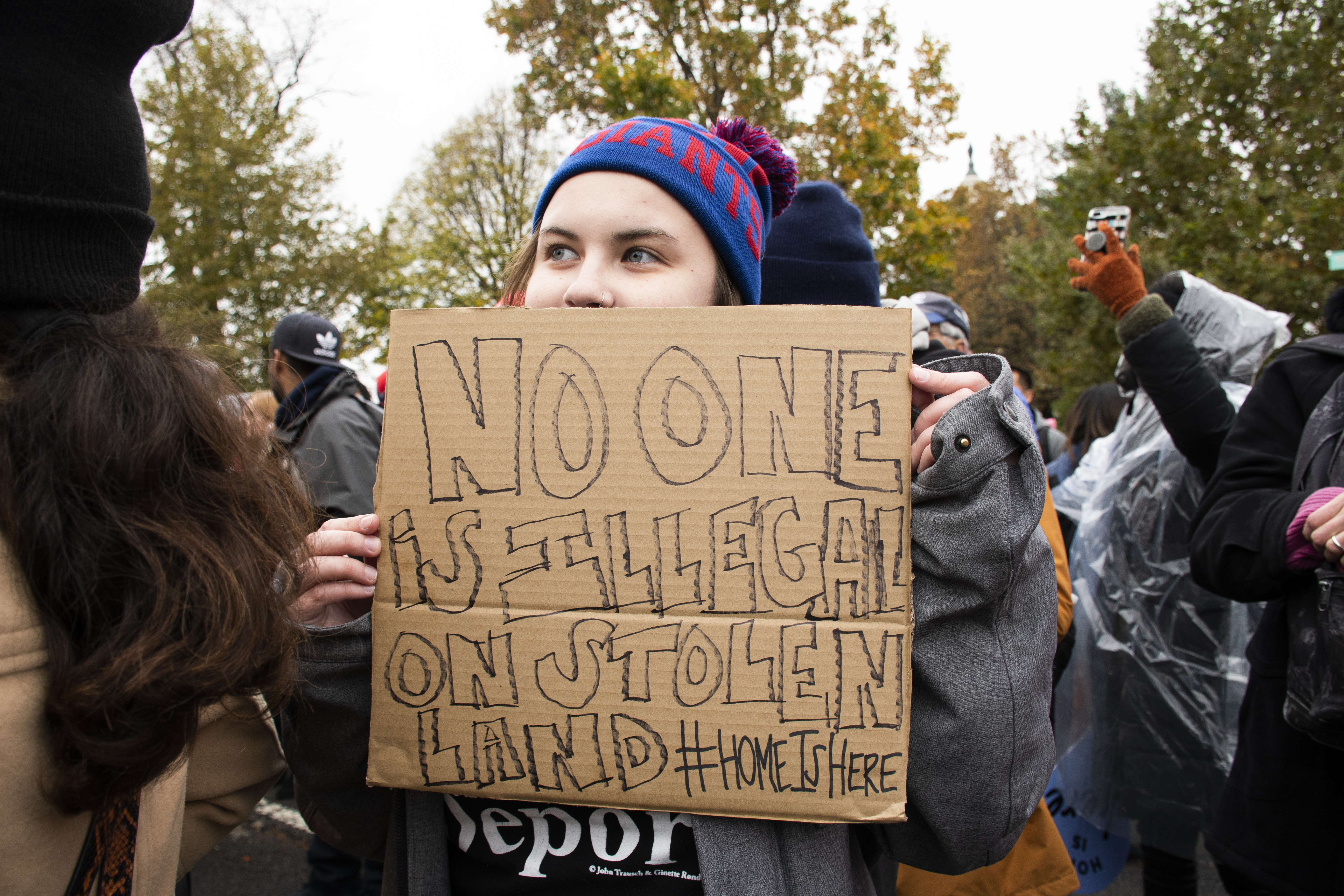 A woman peers over a cardboard sign that reads: No one is illegal on stolen land #HomeIsHere