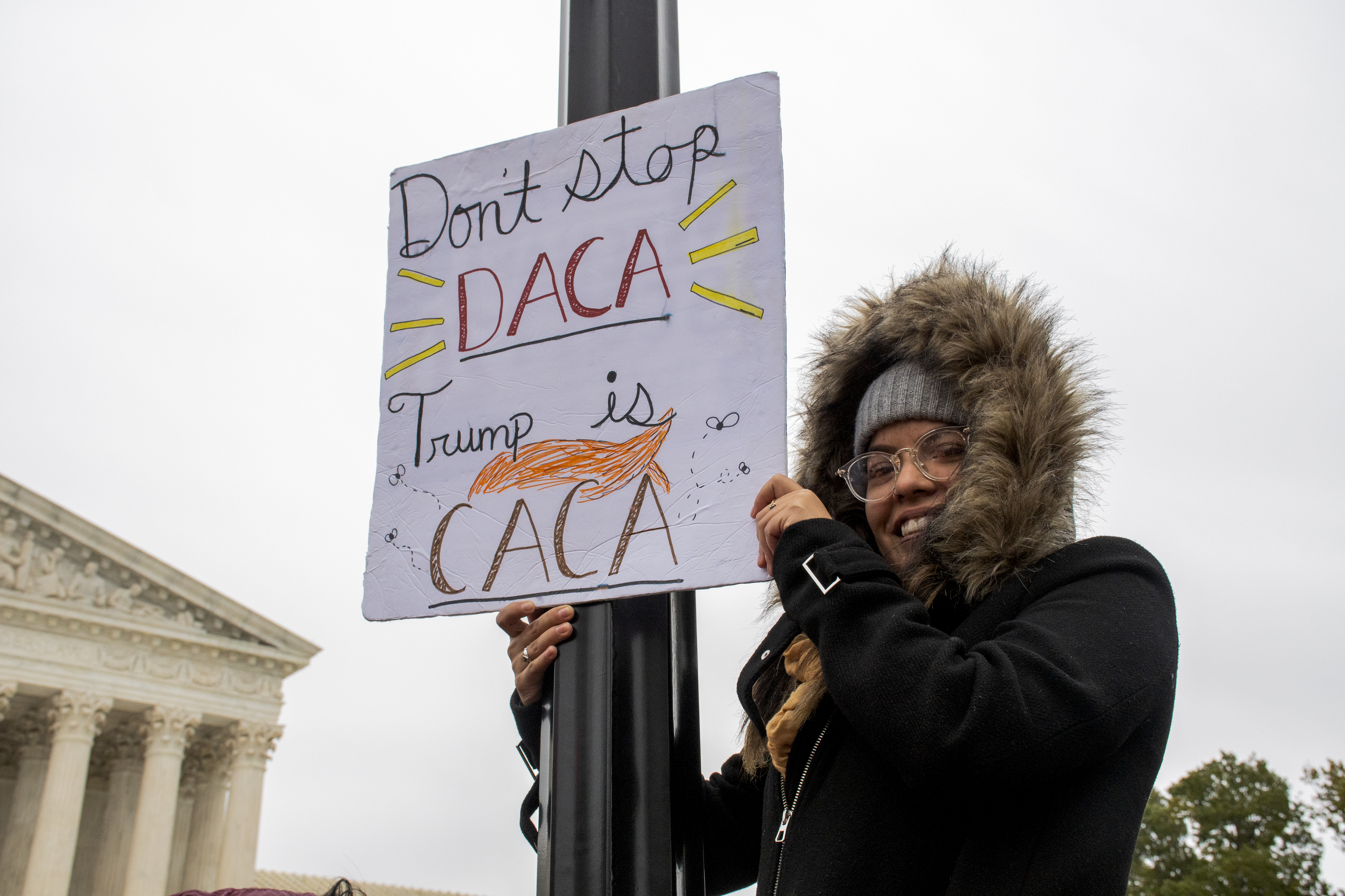 Woman in a winter coat balanced on a pole in front of the Supreme Court holds up a sign that reads: Don't stop DACA, Trump is CACA