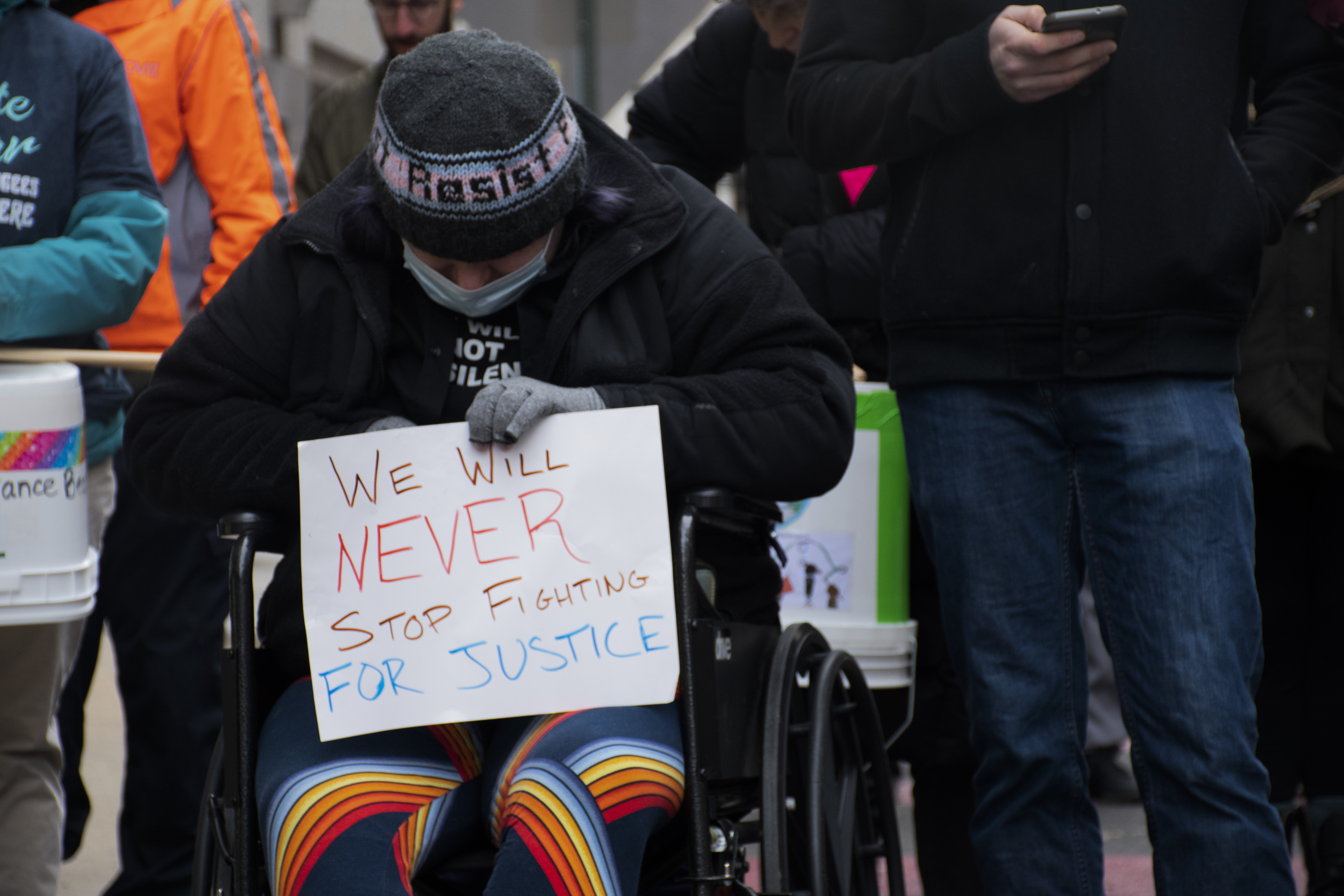 Woman in a wheelchair with her head bent holds up a sign that says: We will never stop fighting for justice